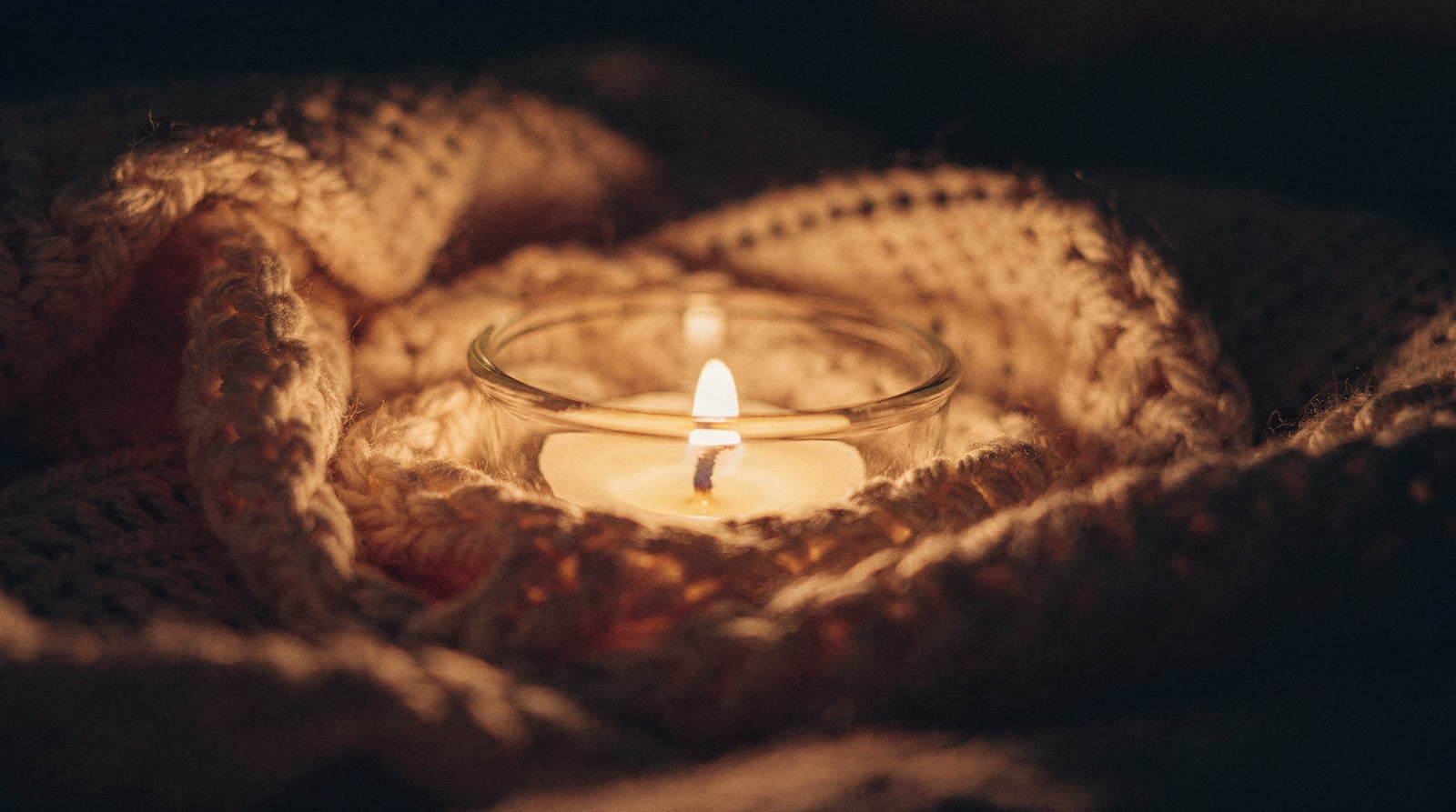A lit candle nestled in a soft cream knitted blanket in warm low light