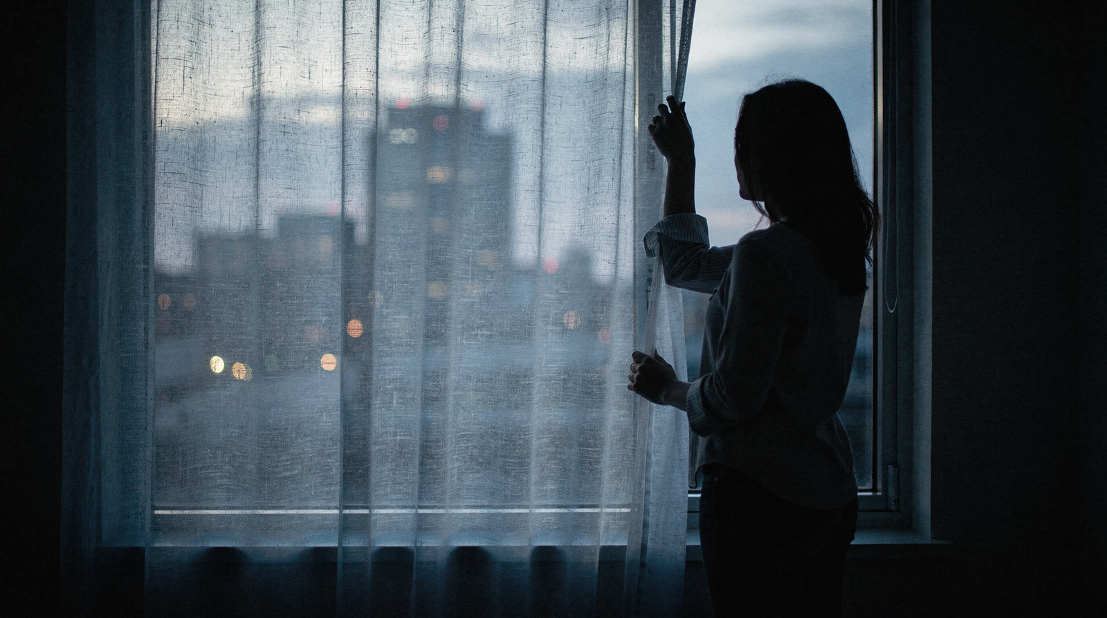 Silhouette of a woman standing at a window at dusk in cool blue light