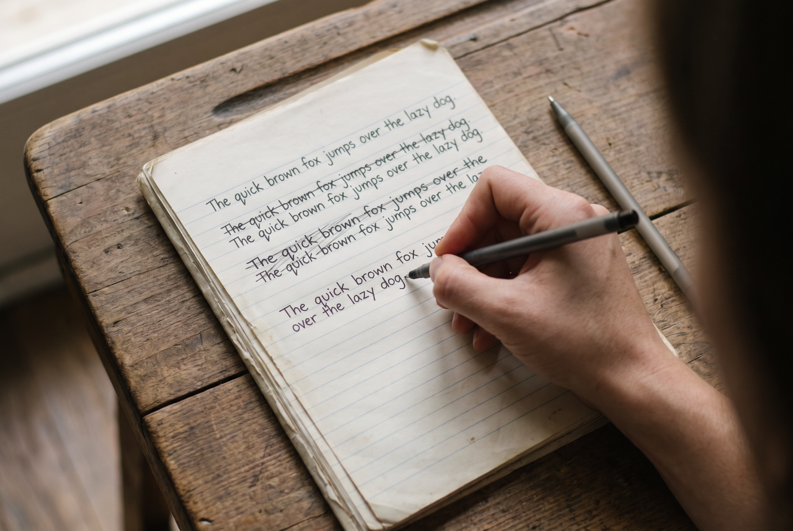Close overhead shot of a hand rewriting the same sentence for the fourth time, with three previous crossed-out attempts visible