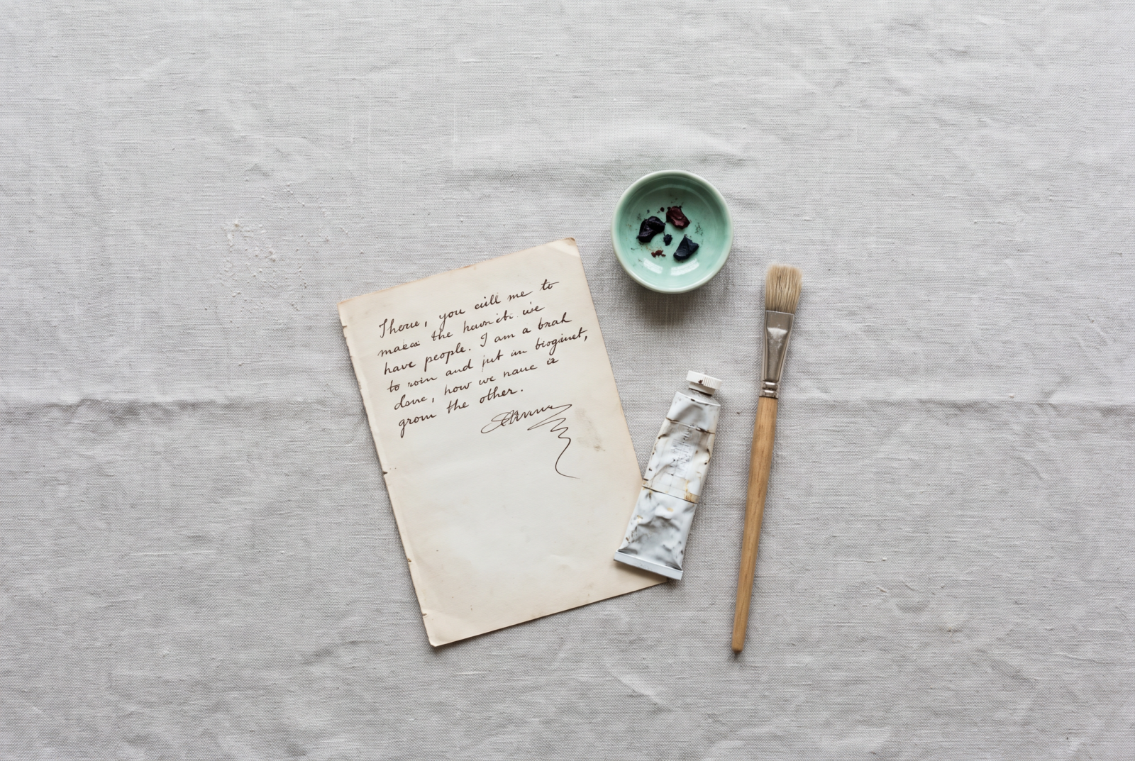 Editorial flat lay of a half-finished manuscript page, dry paintbrush beside untouched paint, quiet stillness and a bit of dust implied