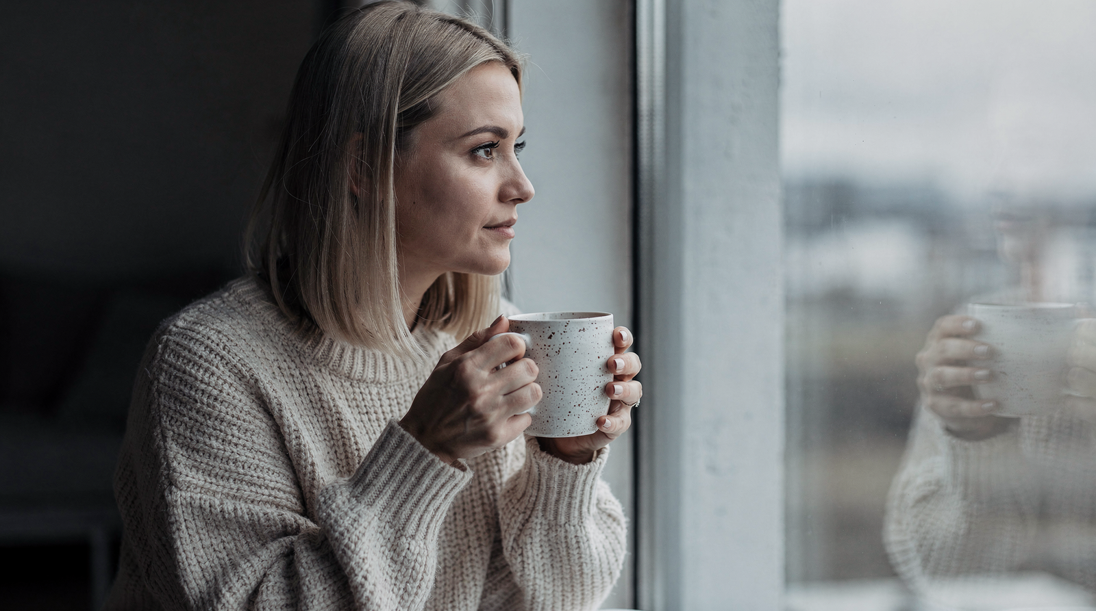 Woman in a cream knit sweater sitting by a window holding a mug
